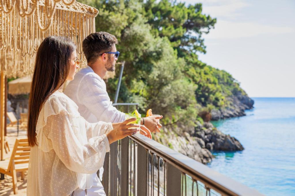 Pāris, baudot mierīgu skatu uz jūru no terases, tur rokās kokteiļus un izskatās relaksēti. Apkārt ir zaļi koki un klusā jūras ūdens virsma.