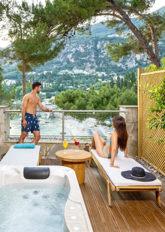 Atpūtas vieta ar skatu uz jūru, kur divi cilvēki relaksējas, viens atrodas pie baseina, bet otrs sēž uz sauļošanās krēsla. Apkārt ir zaļa daba un kalnu ainava.