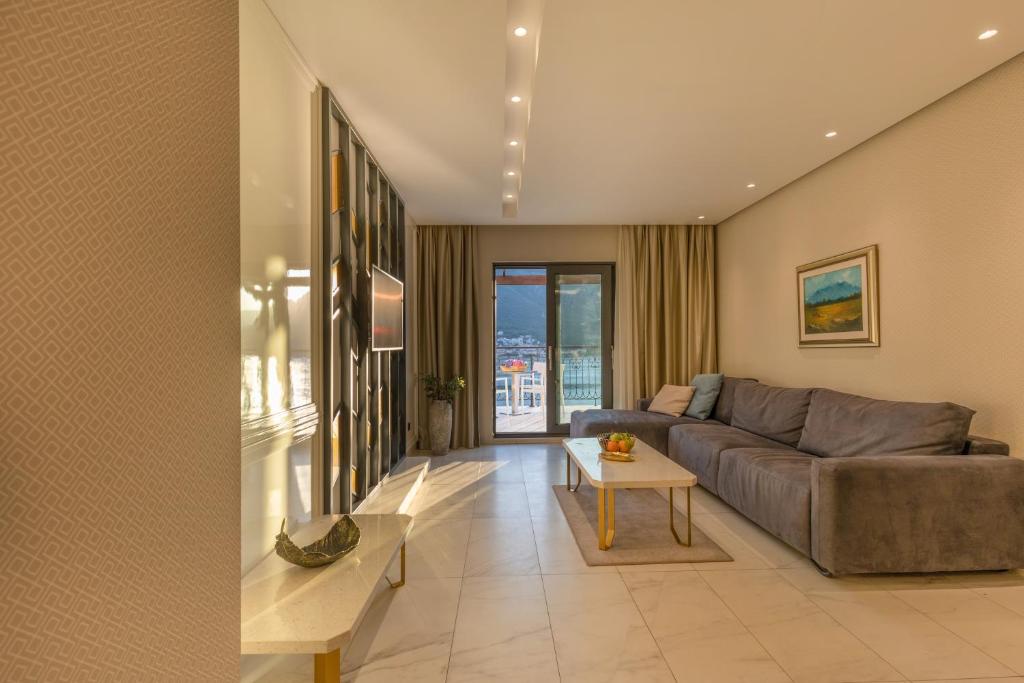 Mūsdienīgs viesistabas interjers ar pelēku dīvānu, koka galdu un skatu uz ārpusi. Telpā ir gaismas un eleganta apdare.