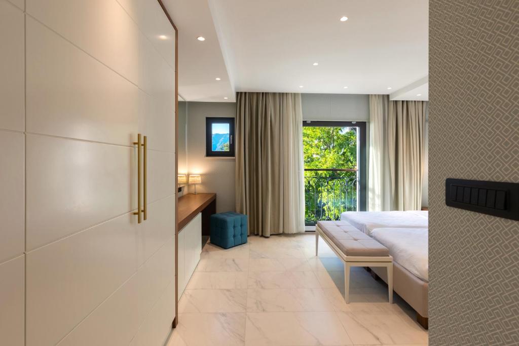 Mūsdienīga viesnīcas numuriņa interjers ar gaišām sienām un dabīgu apgaismojumu, redzami lieli logi un balkons ar skatu uz dārzu. Eleganta mēbeles un neitrālas krāsas rada relaksējošu atmosfēru.