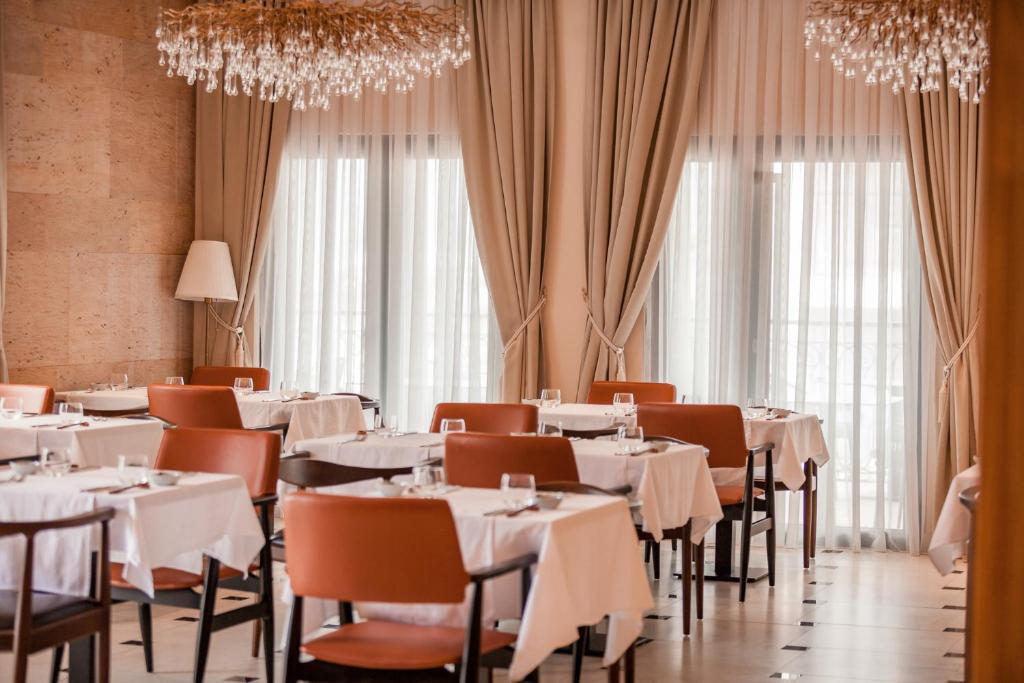 Elegants restorāns ar baltām galdautām un siltām krēslu tonām, apgaismots ar izsmalcinātu lustru. Lielas logu aizkari ļauj ienākt dabiskajai gaismai.