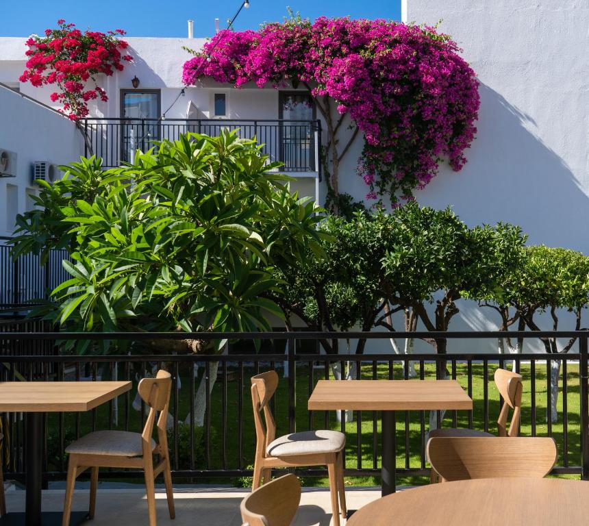 Dārza terase ar koka galdiem un krēsliem, ko ieskauj zaļumi un krāsaini ziedi, radot relaksējošu atmosfēru. Ziedu pušķi pie mājas fasādes piešķir dzīvīgumu ainavai.