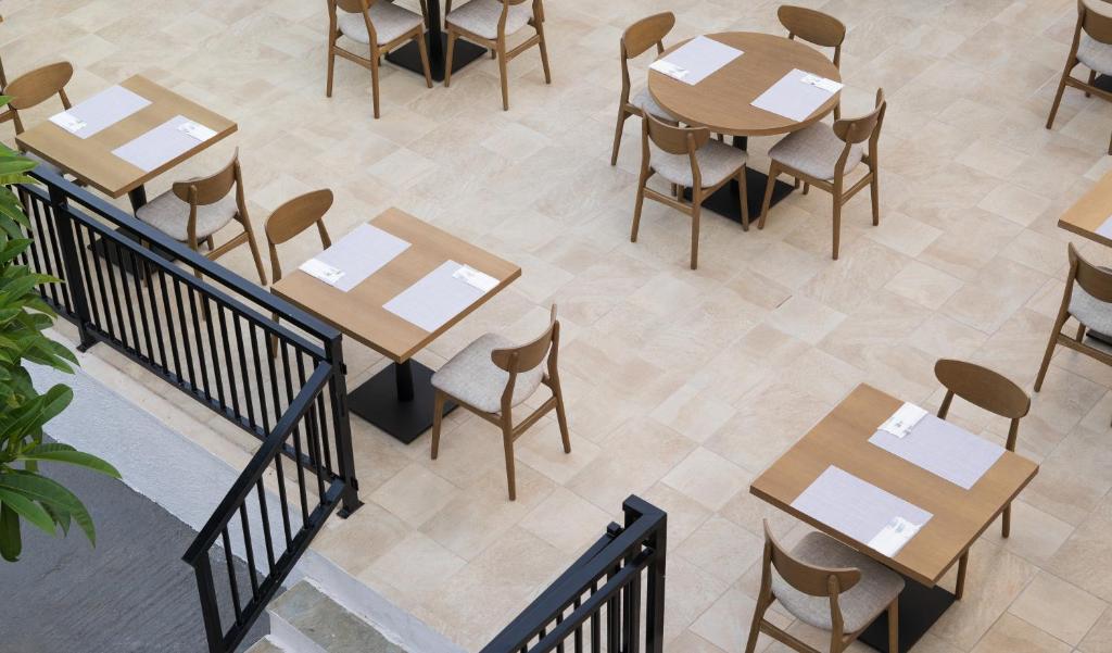 Restorāna interjers ar koka galdiem un krēsliem, kas izvietoti uz gaišām flīzēm. Redzami galdi ar ēdienkarti uz tiem un dārza auga daļa attēla malā.