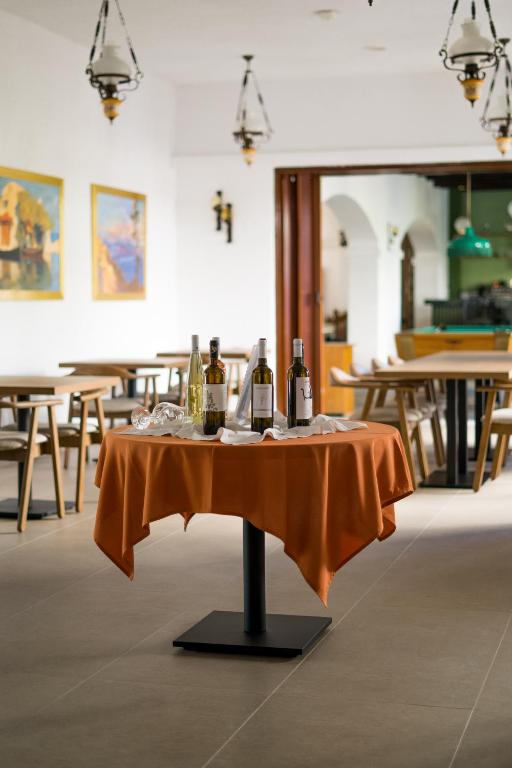 Galda ar oranžu galdautu un vīna pudelēm restorānā, apkārt redzami koka galdi un gleznas uz sienām.