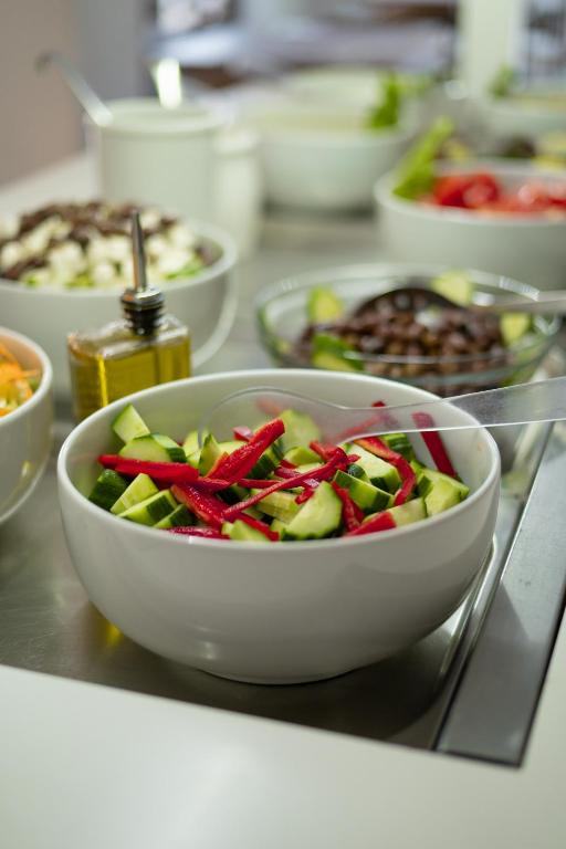 Svaiga dārzeņu salāti, kas satur gurķus un paprika, pasniegti baltā bļodā, apņemti ar citiem dārzeņiem un eļļu uz galda.