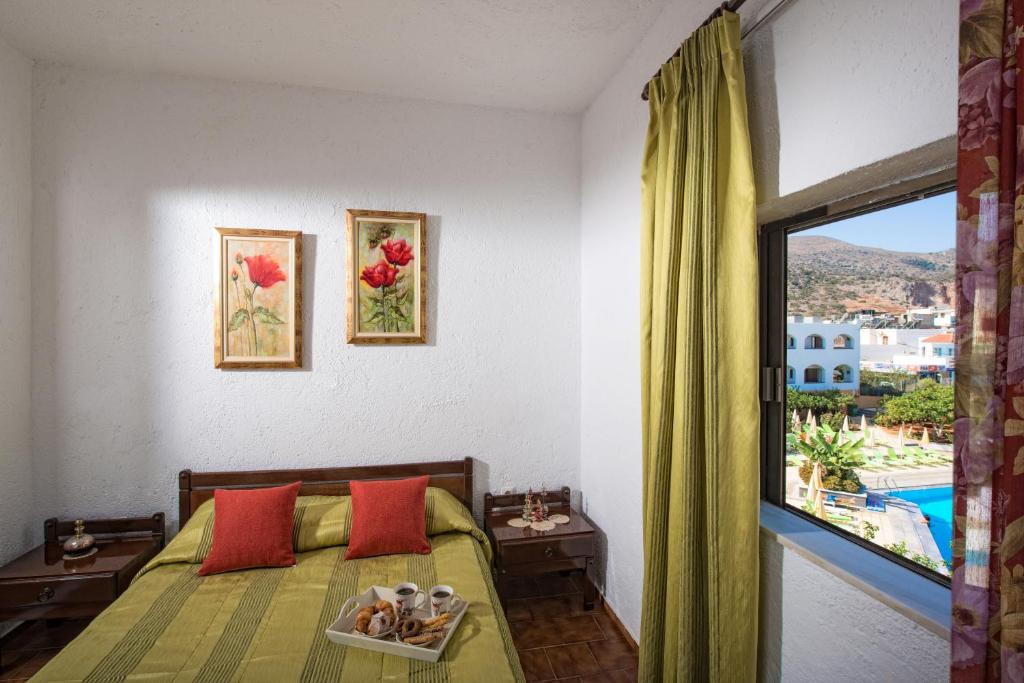 Mūsdienīga guļamistaba ar zaļu gultas veļu, sarkaniem spilveniem un gleznām uz sienas. Logā redzams skats uz apkārtni un baseinu.