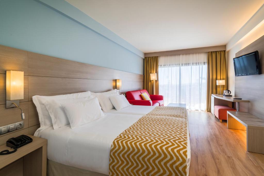 Mūsdienīga viesnīcas numura iekārtojums ar divām gultām, sarkanu dīvānu un skatu uz ārpusi caur lieliem logiem. Interjers ir gaišs un viesmīlīgs, radot komfortablu atmosfēru.