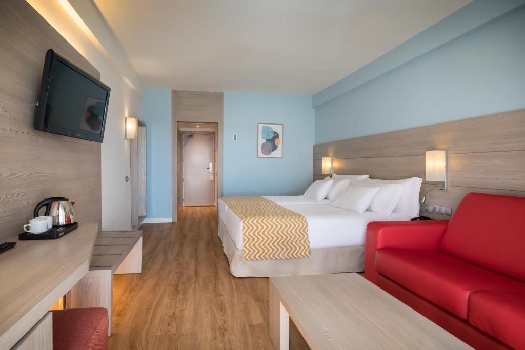 Moderns viesnīcas numurs ar divām gultām, sarkanu dīvānu un televizoru uz koka sienas. Telpā ir gaišas krāsas, koka grīda un minimālistisks dizains ar mākslas darbu uz sienas.