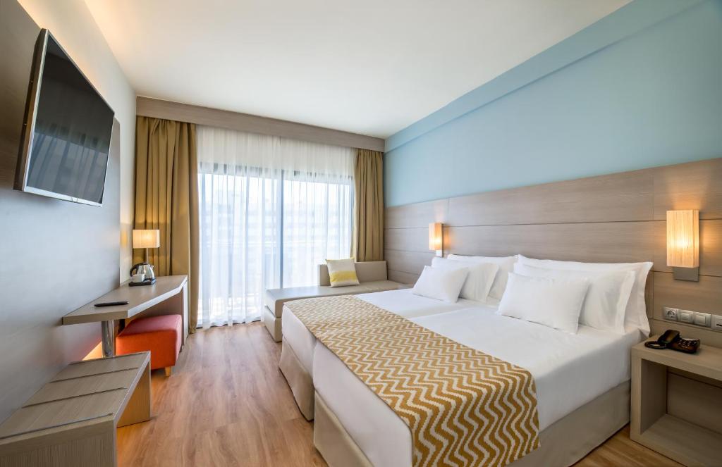 Mūsdienīgs viesnīcas numurs ar divām gultām, televizoru un daļēji atvērtu terasi. Interjerā dominē gaiši toņi un koka grīdas segums, radot ērtu un patīkamu atmosfēru.