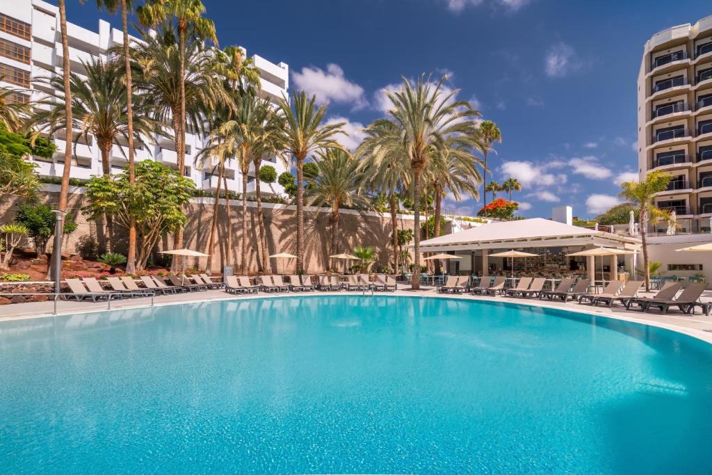 Liela, skaidra peldbaseins, ko ieskauj palmas un sauļošanās krēsli, atrodas viesnīcas teritorijā ar modernu arhitektūru un zilu debesu fonu.