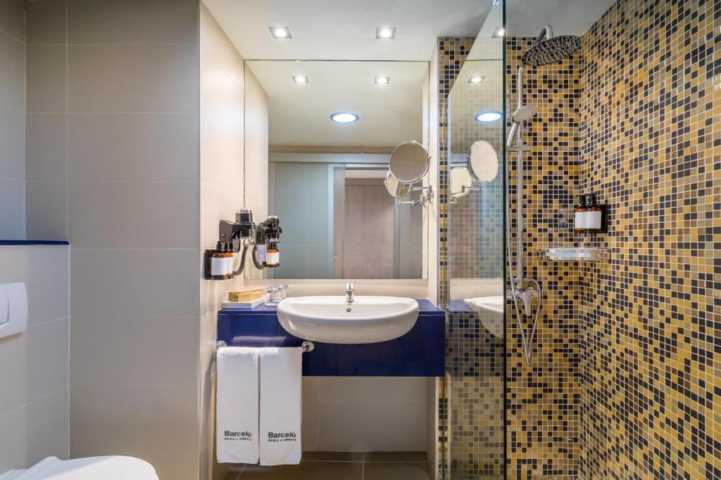 Mūsdienīgs vannasistabas interjers ar mozaīkas sienām, stikla dušas kabīni un elegantu izlietni. Vannas istabā ir arī spoguļi un higiēnas piederumi.