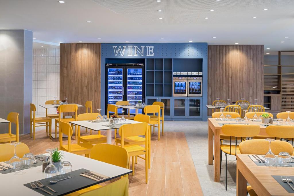 Mūsdienīgs restorāns ar dzeltenām krēsliem un elegantiem galdiem, kurā redzams vīna stends un atvērtas vitrīnas. Interjers apvieno koka un mūsdienīgus elementus, radot patīkamu atmosfēru.