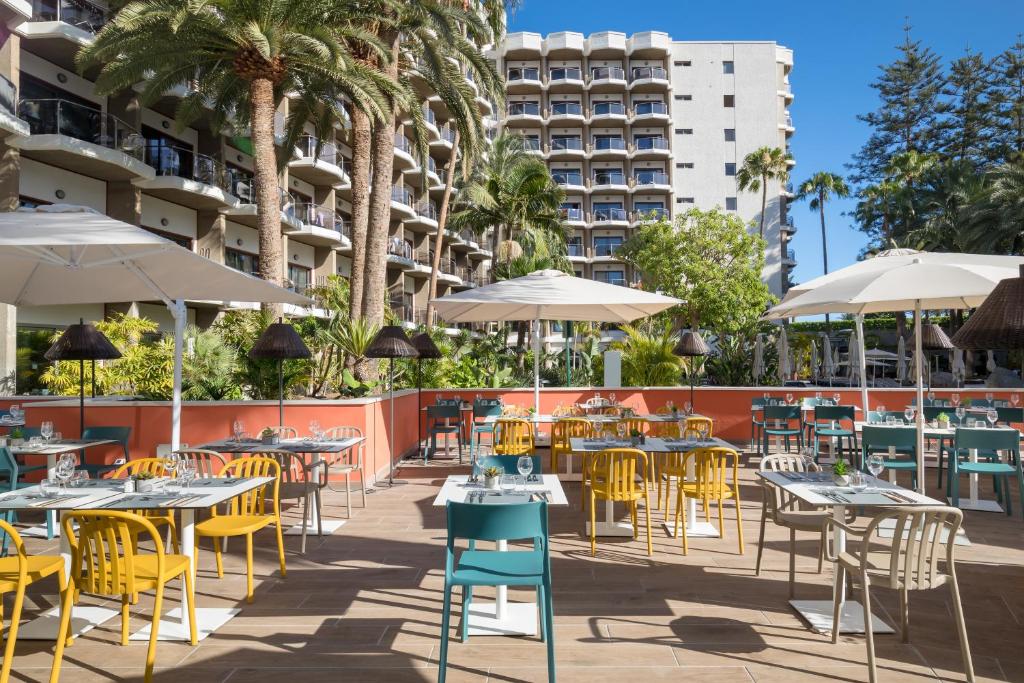 Āra restorāna zona ar krāsainiem galdiem un krēsliem, apzaļumota ar palmām un sauļošanās lietussargiem. Fonā redzams viesnīcas ēka ar balkoniem.