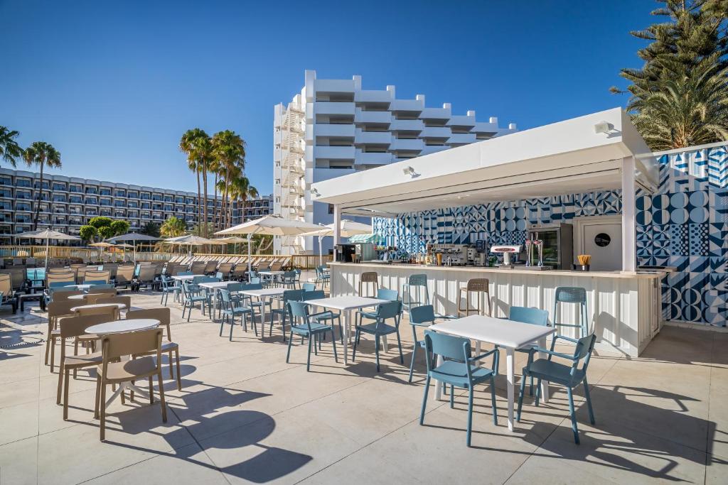 Peldbaseina zona ar modernu bāru un ēdināšanas vietu, kur ir gaiši galdi un krēsli, palmām un viesnīcas ēku fonā. Ideāla vieta atpūtai un relaksācijai.