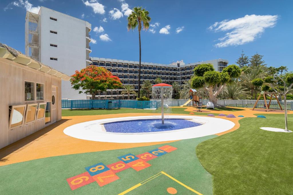 Bērnu rotaļu laukums ar baseinu un krāšņām ainavām, kas atrodas viesnīcas pagalmā. Iespējami aktivitāšu elementi, piemēram, šūpoles un slidkalniņš, ko ieskauj zaļumi un ziedi.