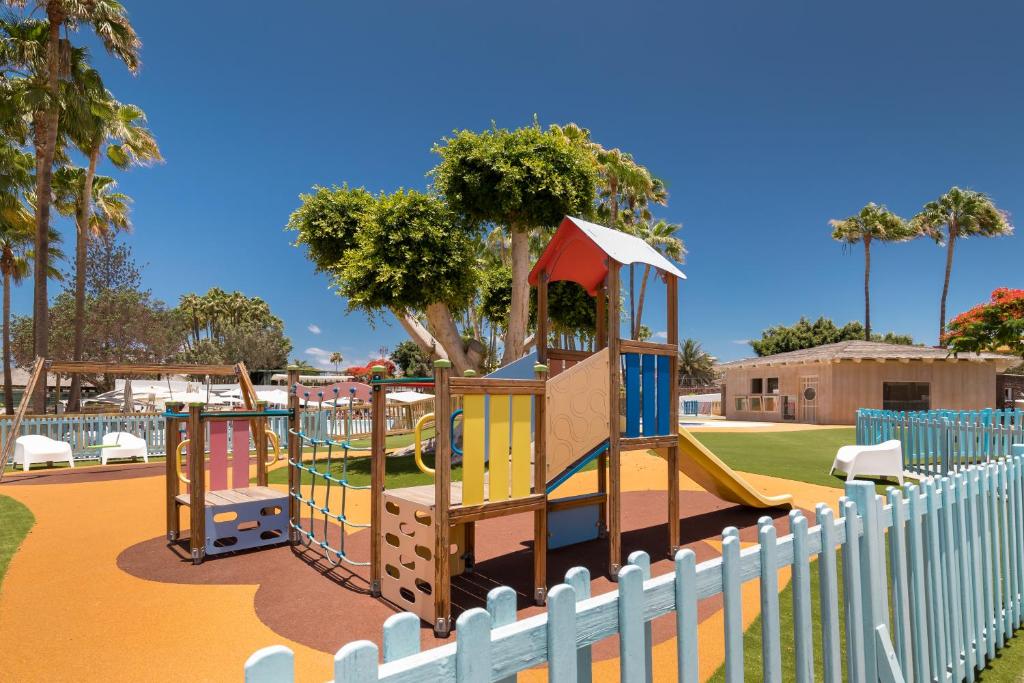 Bērnu rotaļu laukums ar krāsainiem spēļu elementiem un šūpolēm, apkārt zaļojošām palmām un skaidru zilo debesu fonu.