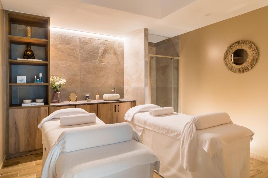 Mūsdienīgs spa telpa ar divām masāžas galdiem, dekorēta ar neitrālām krāsām un relaksējošu apgaismojumu. Uz plauktiem redzami skaistumkopšanas produkti un ziedi, radot mierīgu atmosfēru.