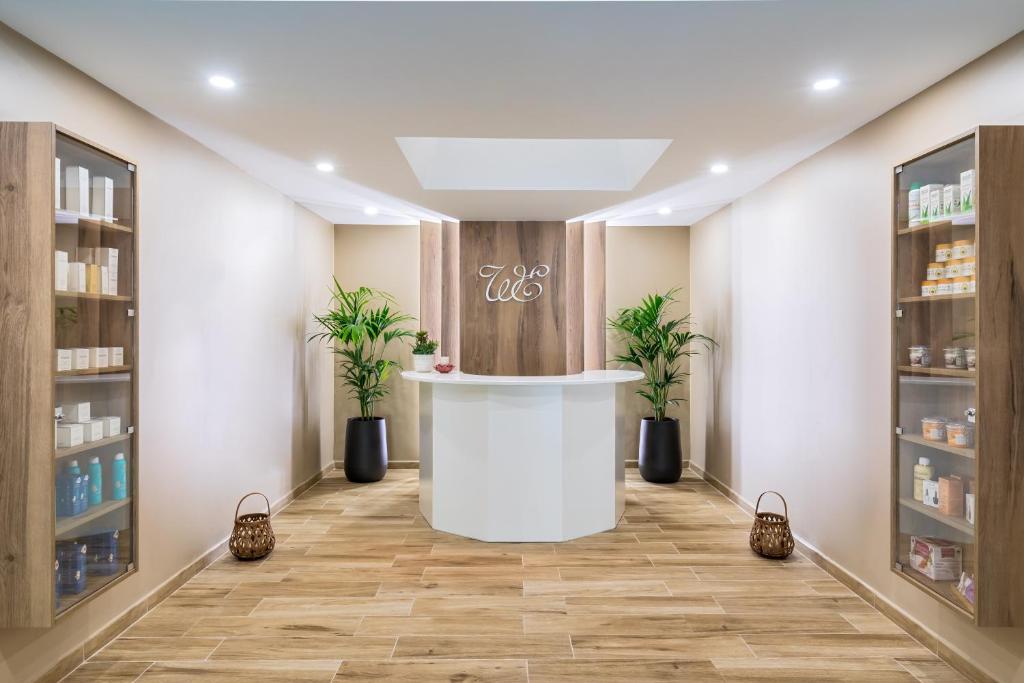 Moderns un stilīgs skaistumkopšanas salons ar baltu reģistratūru un elegantiem koka skapjiem, kas piepildīti ar kopšanas produktiem. Telpa ir gaiša un aicinoša ar zaļām augiem abās pusēs.