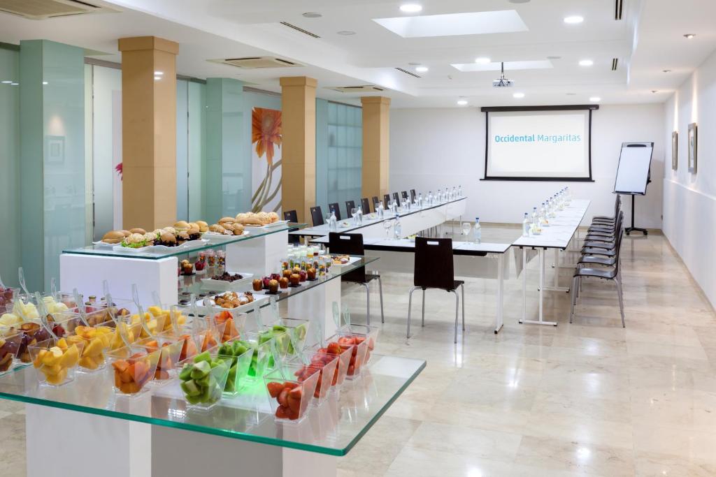Konferenču telpa ar garām galdiem, kur sagatavots uzkodu galds ar augļiem un maizītēm, ideāli piemērota sanāksmēm un pasākumiem. Uz sienas ir projektors ar logo 