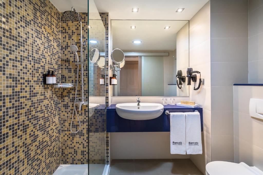 Mūsdienīgs vannas istabas interjers ar mozaīkas flīzēm, dušas kabīni un plašu spoguli. Vannas istabā redzami higiēnas piederumi un eleganta, minimāla dizaina jaucējkrāns.