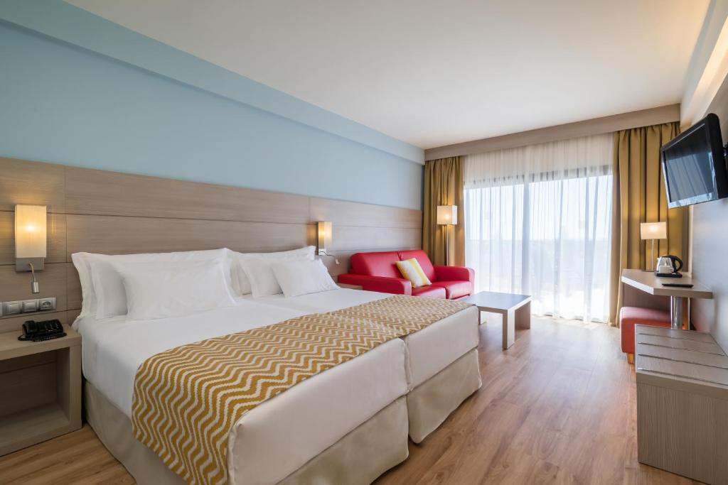 Mūsdienīga viesnīcas numura iekārtojums ar divām gultām, sarkanu dīvānu un logu, caur kuru ieplūst dabiska gaisma. Numurā ir televizors un galdiņš ar krēslu.