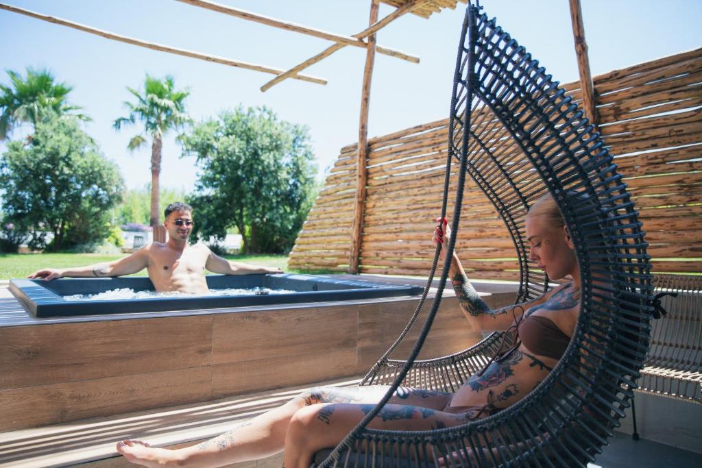 Atpūtas brīvdienas pie baseina, kur divi cilvēki izbauda sauli un mierīgu atmosfēru. Sieviete šūpojas modernā šūpuļkrēslā, bet vīrietis relaksējas baseinā.