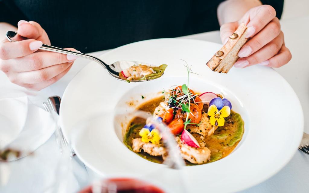 Gourmet maltīte ar ziediem, dārzeņiem un garšvielām, ko pasniedz elegantā traukā. Rokas tur karoti un maizes gabalu, radot izsmalcinātu gastronomijas pieredzi.