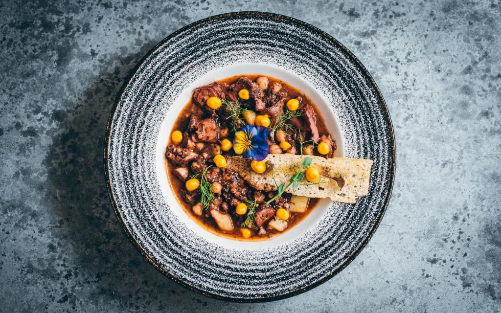 Skaisti pasniegta zupa ar gaļu, dārzeņiem un dekoratīvām ziedlapiņām, uz eleganta šķīvja ar melnu un baltu dizainu.