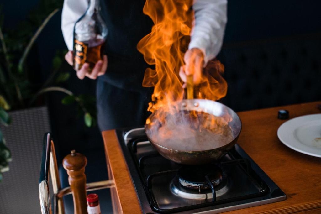 Šefpavārs sagatavo ēdienu, izmantojot uguni, kas iznāk no pannas. Attēls parāda dinamisku gatavošanas procesu, kurā tiek izmantots alkohols.