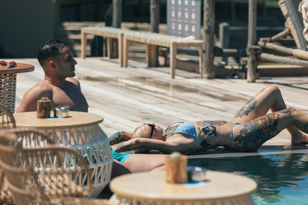 Divi cilvēki atpūšas pie baseina, baudot sauli. Viens no viņiem ir ar tetovējumiem un bikini, bet otrs - ar īsām biksēm. Apkārt ir moderni mēbeļu gabali un relaksējoša atmosfēra.