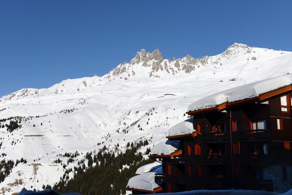 Ziemas kalnu ainava ar sniega klātiem pakalniem un koka ēkām, kas atrodas priekšplānā. Skaidrs zils debesis piešķir attēlam mierīgu un svaigu noskaņu.