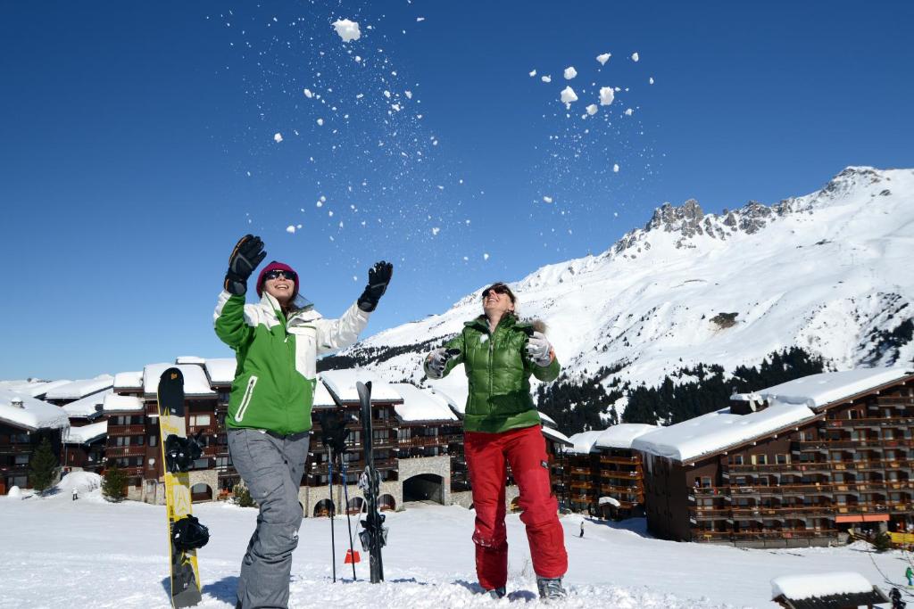Divas cilvēku grupas izbauda ziemas aktivitātes sniegā, spēlējoties un mestot sniega kamolus, kamēr fonā redzams kalns un sniegoti kūrorti.