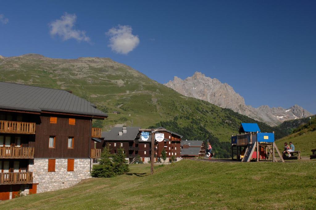 Kalnu ainava ar modernām mājām un bērnu spēļu laukumu, kas atrodas zaļā pļavā. Fons ir majestātiskas kalnu virsotnes un skaidrs debesis.