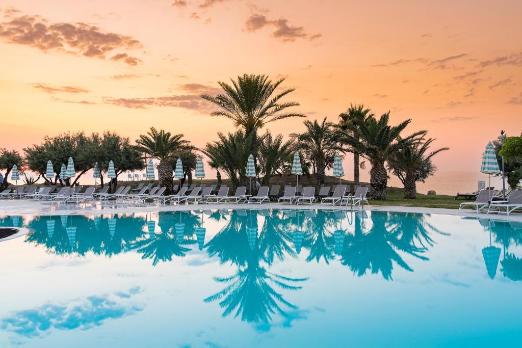 Miera osta ar baseinu un palmu siluetu, saules rietumu gaismā. Skats uz mierīgu ūdeni, kas atspoguļo debesu krāsas.