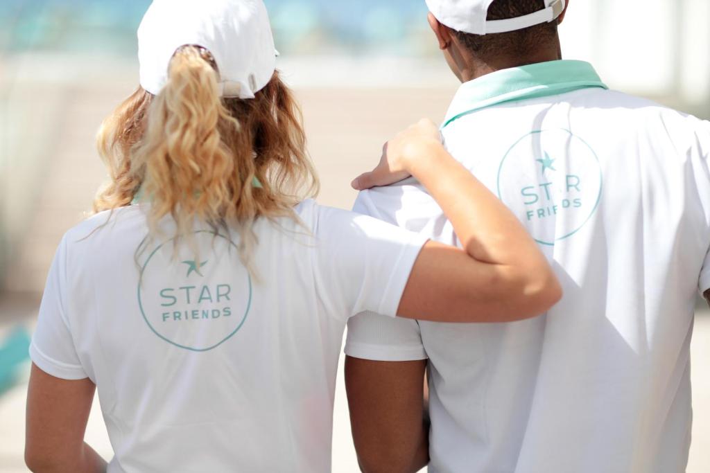 Divi cilvēki, tērpti baltās polo krekliņos ar logo "STAR FRIENDS", stāv kopā, viens otram atbalstot plecus. Attēls izsaka draudzību un komandas garu.