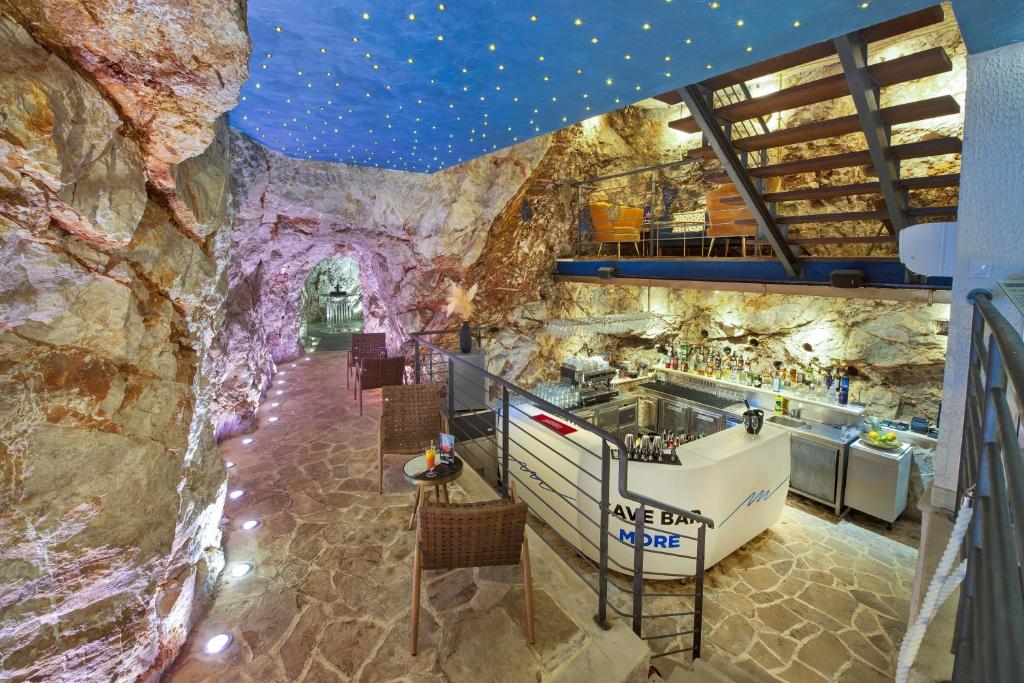 Unikāls klinšu bārs ar stilīgu interjeru, kas apvieno dabas elementus un modernu dizainu. Telpā redzamas akmens sienas un apgaismojums, kas rada noslēpumainu atmosfēru.