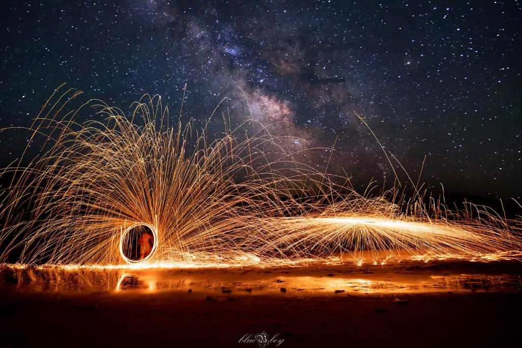 Degoši stieņi veido gaismas efektus, kamēr debesīs redzama Milky Way. Šī nakts ainava iemūžina brīnišķīgu kombināciju starp uguni un zvaigznēm.