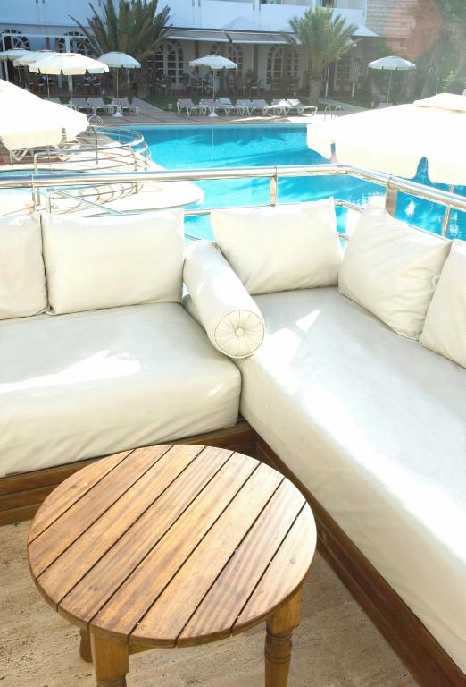 Mūsdienīgs atpūtas stūrītis ar baltām dīvānu spilveniem un koka galdiņu, skatoties uz baseinu un sauļošanās krēsliem. Ideāla vieta relaksācijai un atpūtai.