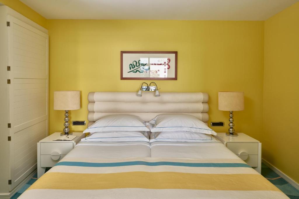 Mūsdienīga guļamistaba ar dzeltenām sienām, ērtu gultu un stilīgām lampām. Interjers ir gaišs un viesmīlīgs, radot relaksējošu atmosfēru.
