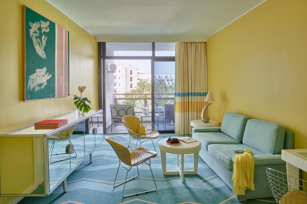 Mūsdienīga viesnīcas numura interjers ar gaiši dzeltenām sienām, zilā paklāja un mēbeļu kombināciju, kā arī modernu mākslas darbu pie sienas. Iekārtojums rada svaigu un komfortablu atmosfēru.