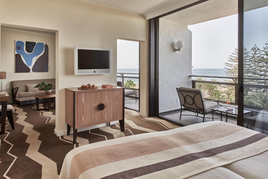 Mūsdienīga viesnīcas istaba ar jūras skatu, iekārtota ar elegantiem mēbeļu elementiem un mākslas darbu uz sienas. Gaismas tonis un plašās logu konstrukcijas nodrošina patīkamu un relaksējošu atmosfēru.