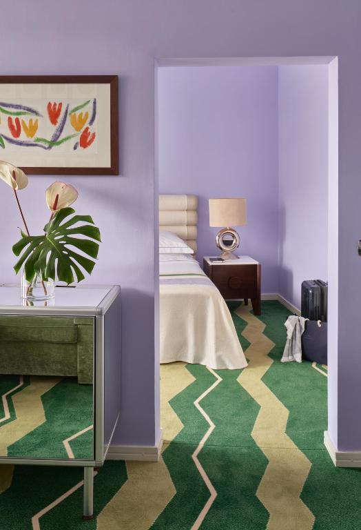 Mūsdienīgs interjers ar violetām sienām, zaļu paklāju ar viļņotu rakstu un ziedu kompozīciju uz galda, kas vērš uzmanību uz guļamistabas daļu.