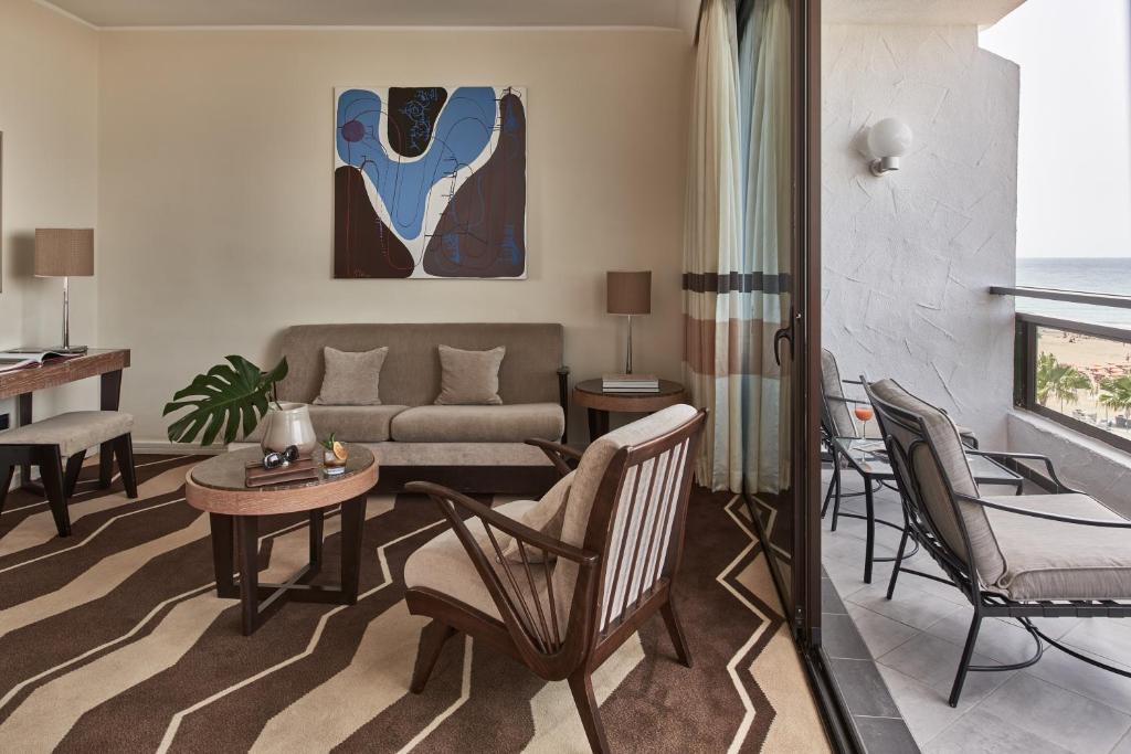 Mūsdienīgs viesnīcas numurs ar jūras skatu, ērtiem mēbeļiem un mākslas darbu uz sienas. Telpā ir dīvāns, krēsli un galds, kā arī balkons ar atpūtas krēsliem.