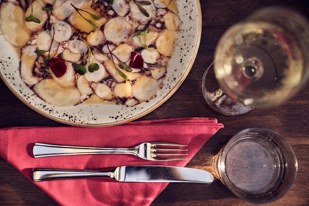 Grezns ēdiens uz šķīvja ar garnelēm un dārzeņiem, papildināts ar vīna glāzi un galda piederumiem uz sarkanas salvetes.