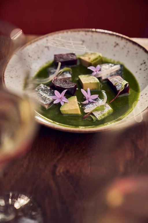 Grezna šķīvja kompozīcija ar krāsainiem dārzeņu gabaliņiem un ziediem, pasniegta zaļā mērces bāzē.