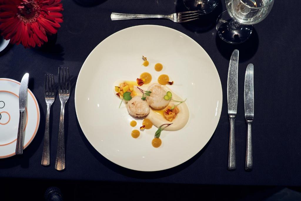 Grezns ēdiens uz baltas šķīvja ar dekoratīviem elementiem un krāsainām mērču pilēm, novietots uz melna galdauta. Ap galdu ir eleganti galda piederumi un dzērienu glāzes.