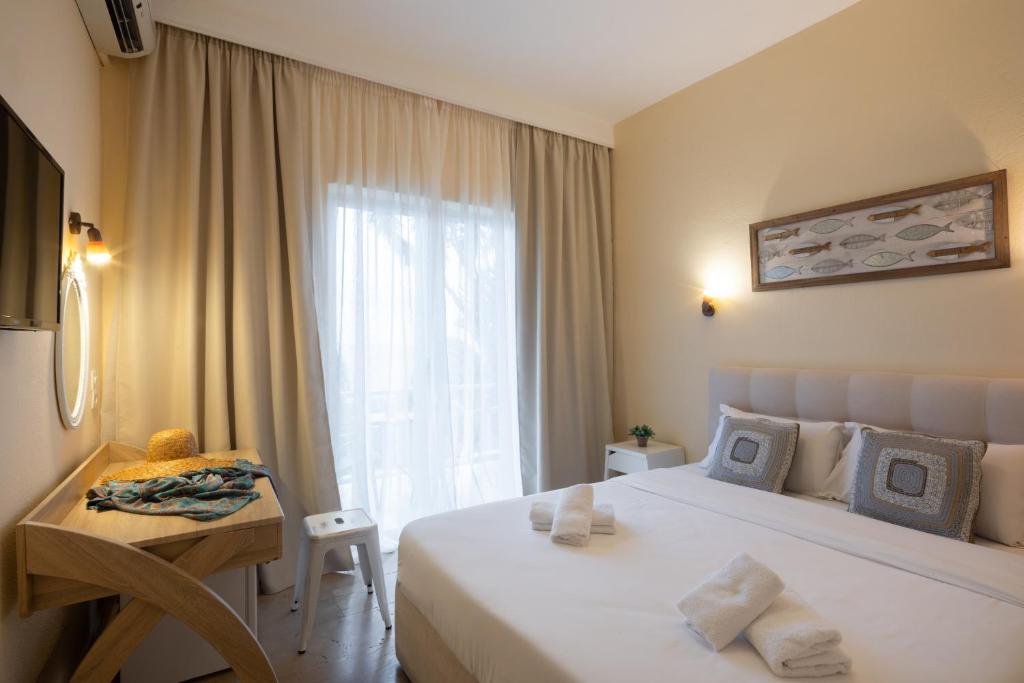 Gaiša un mūsdienīga viesnīcas istaba ar divguļamo gultu, dīvānu un koka galdu. Pie loga redzamas vieglas aizkari, bet uz galda novietoti aksesuāri un dvieļi.