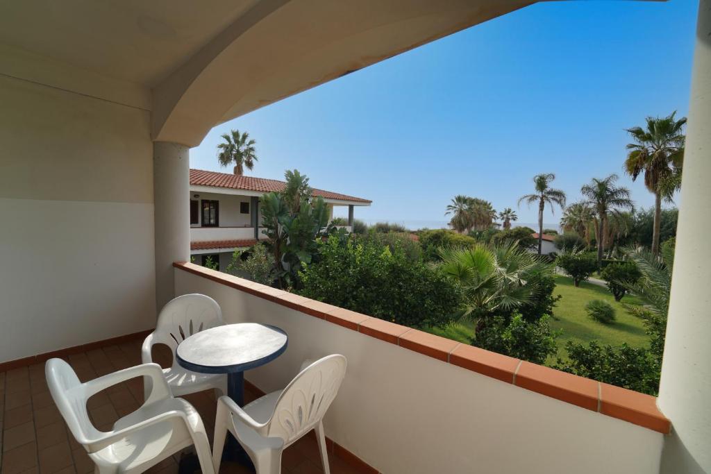 Balkons ar galdu un krēsliem, redzams skats uz dārzu un palmām, skaidra debess.