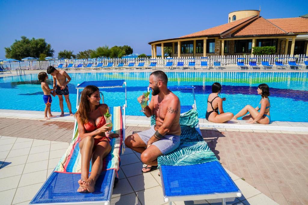 Vasaras brīvdienu atmosfēra pie baseina, kur pāris izbauda kokteiļus, kamēr bērni spēlējas tuvumā, un citas personas atpūšas saulē.