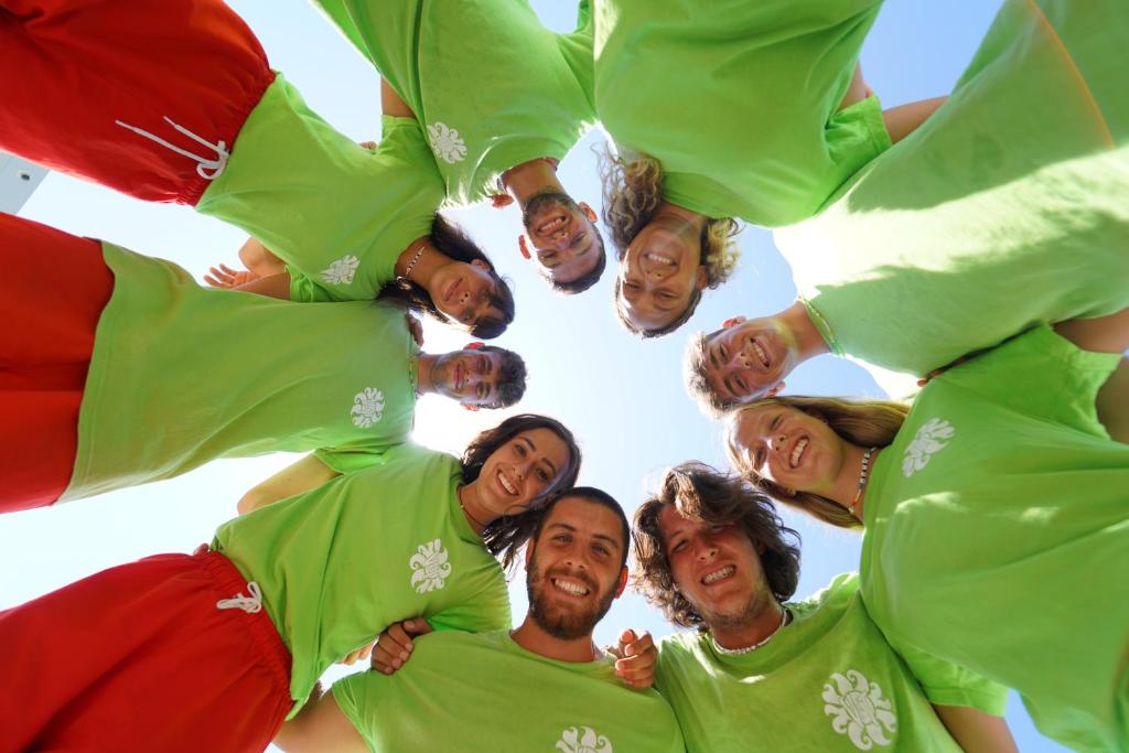 Grupas jauniešu fotogrāfija, kuri valkā spilgti zaļas t-kreklus un sarkanas bikses, stāv aplī ar paceltām rokām, priecīgā atmosfērā zem saules.
